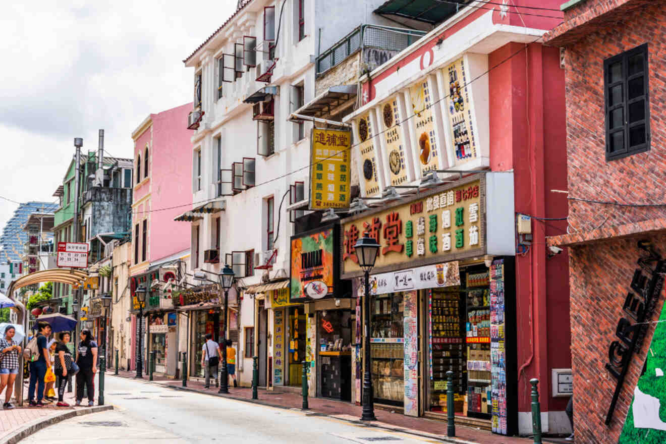 Colorful street in Taipa, Macau, with traditional low-rise buildings, various shop signs in Chinese, and a few pedestrians walking along the quiet road.