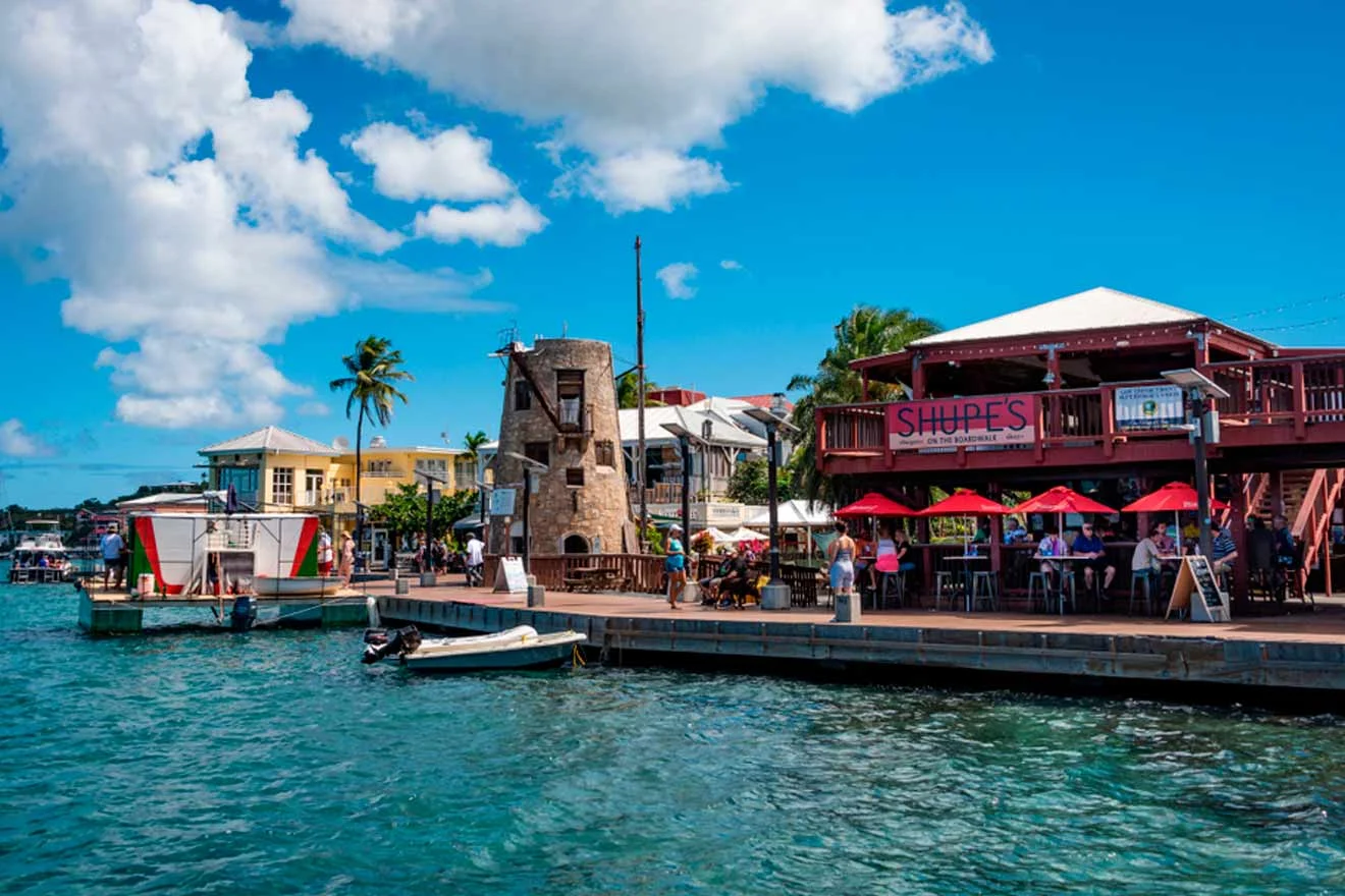 A waterfront scene with a dockside restaurant featuring red umbrellas and patrons dining. Boats are moored along the water, and buildings with palm trees are in the background under a partly cloudy sky.