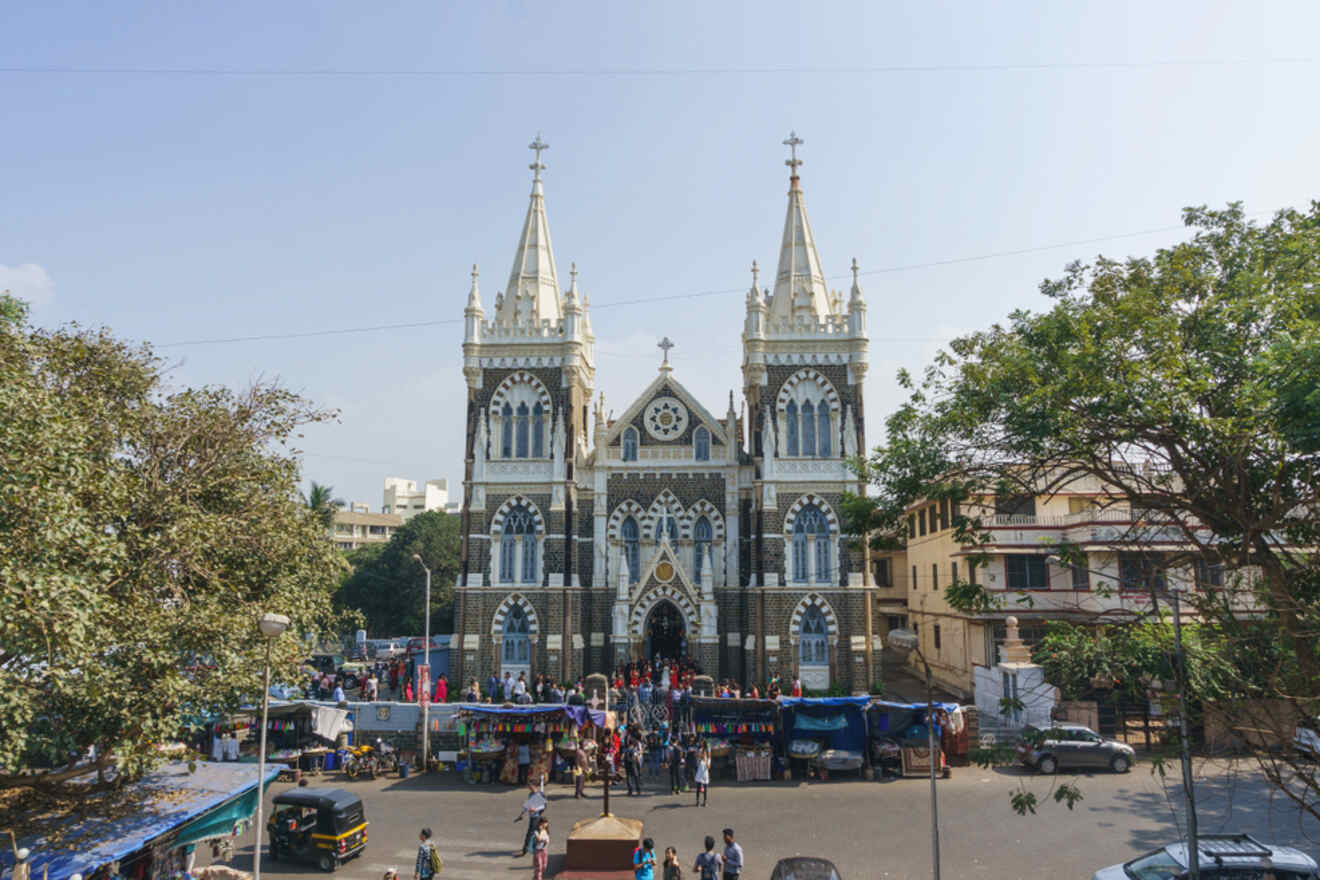 A large Gothic-style cathedral with two tall spires in a bustling square surrounded by trees, people, and market stalls.