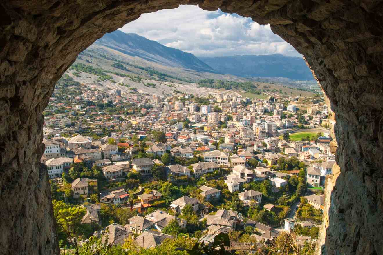 A panoramic view of Gjirokastër, Albania, framed by a stone archway, showcasing traditional stone houses spread across the valley with mountains in the background