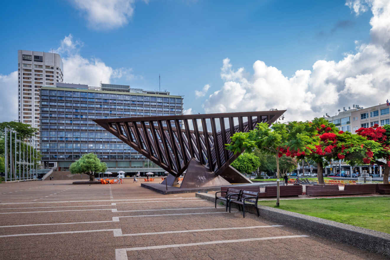 An expansive plaza with a modern triangular monument in the center, surrounded by trees with bright red flowers, benches, and a large government building in the background.