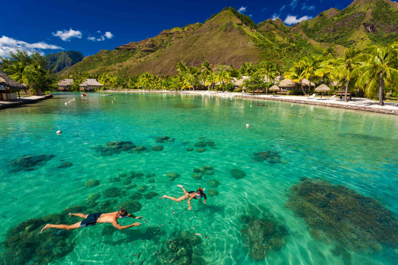 Two people snorkel in clear turquoise water near coral formations, with lush, green mountains and overwater bungalows in the background under a bright blue sky.