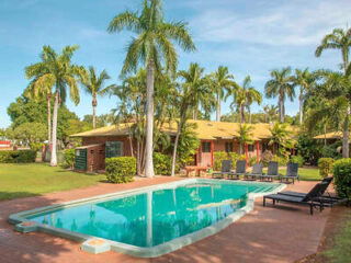 A backyard with a swimming pool surrounded by paved flooring, lounge chairs, and palm trees, alongside a one-story house with a yellow roof.