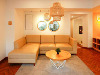 A modern living room with a beige sectional sofa, a round wooden coffee table with a plant, a gray rug, and decorative mirrors on the wall. A woven pendant light hangs from the ceiling.