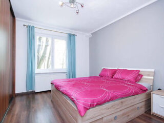 A bedroom with a bed having pink pillows and bedspread, wooden flooring, a window with blue curtains, a gray wall, and a wooden wardrobe.