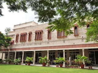 A historic building with arched windows and a balcony, surrounded by green trees and potted plants on a well-maintained lawn.