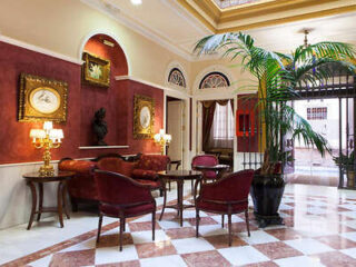 A hotel lobby with red chairs and tables, palm plant, framed artwork on red and cream walls, and a checkered floor.