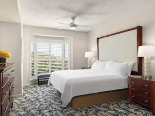 A modern bedroom with a large bed, two bedside tables with lamps, a dresser, a ceiling fan, and a patterned carpet. Sunlight filters through large windows with white shutters.