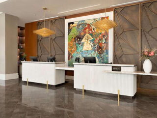 Modern hotel reception area with two white desks, pendant lights, and a large colorful painting on the wall behind.