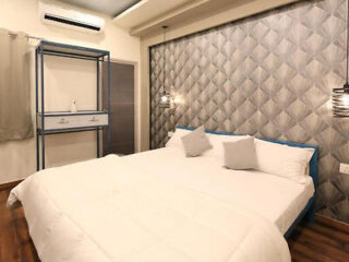 A modern bedroom with a double bed, four pillows, geometric patterned wallpaper, two hanging light fixtures, and a metallic storage rack near the window.