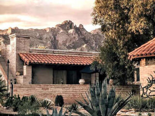 A house with a red-tiled roof, surrounded by desert plants, with a mountain range visible in the background under a cloudy sky.