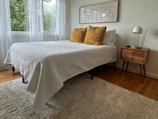 A neatly made bed with a white quilt and two mustard-yellow pillows is placed in a bedroom with white walls, wooden side table, lamp, large window, and soft white rug on the wooden floor.