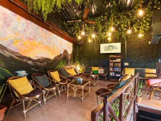 Indoor lounge with wicker furniture, cushions, leafy decor, string lights, a large nature-themed mural, and a small bookshelf. Ceiling fan and television on the wall.