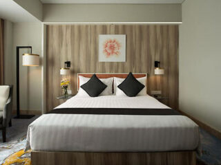 Modern hotel room with a neatly made bed featuring white linens and two black pillows. The bed is against a wooden accent wall with a framed flower picture above. There's a lamp and chair to the left.