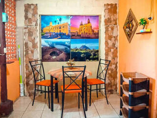 A small dining area with a table, four chairs, and wall decor featuring four colorful landscape pictures. The right wall has a diamond-shaped shelf with small plants, and a black shelving unit stands nearby.