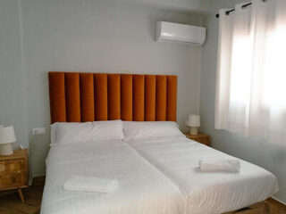 A neatly made bed with white linens and two folded towels, flanked by wooden nightstands with lamps, stands against a wall with an orange upholstered headboard and an air conditioner above.