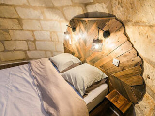 A cozy bedroom features a bed with white linens and a rustic wooden headboard, illuminated by two wall-mounted lamps against a backdrop of stone walls.