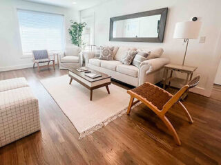 A living room with a beige sofa, patterned cushions, a wooden coffee table, a large mirror, an armchair, and a floor lamp. Interior is decorated with a plant and is well-lit by a window.