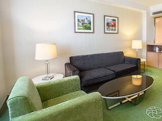 A comfortable hotel living area with a black sofa, green armchair, glass coffee table, and framed wall art