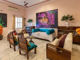 A living room with pastel walls features wooden furniture, a blue cushioned divan, and a vibrant painting of a face. The room is decorated with plants and has marble flooring.