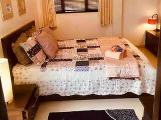 A neatly made bed with a patchwork quilt, several pillows, and folded towels on top. The room includes bedside tables with lamps, light-colored curtains, and a window with blinds.