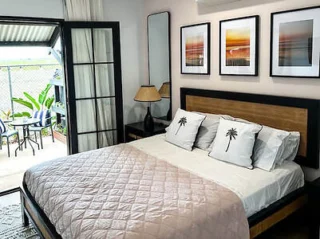 A bedroom featuring a double bed with palm tree patterned pillows, framed artworks above, a side table with a lamp, and an open door leading to a patio with outdoor seating and plants.