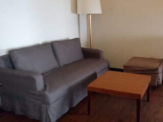 A living room with a gray sofa, wooden coffee table, matching upholstered ottoman, and a floor lamp against a beige wall.