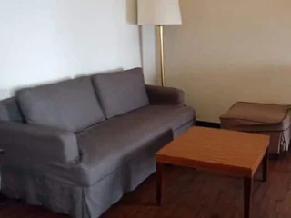 A living room with a gray sofa, wooden coffee table, matching upholstered ottoman, and a floor lamp against a beige wall.