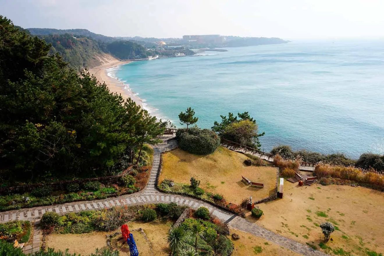 A coastal landscape with a winding path through gardens leading to a sandy beach and blue ocean. Hills with trees are visible in the background. Benches and greenery dot the foreground.