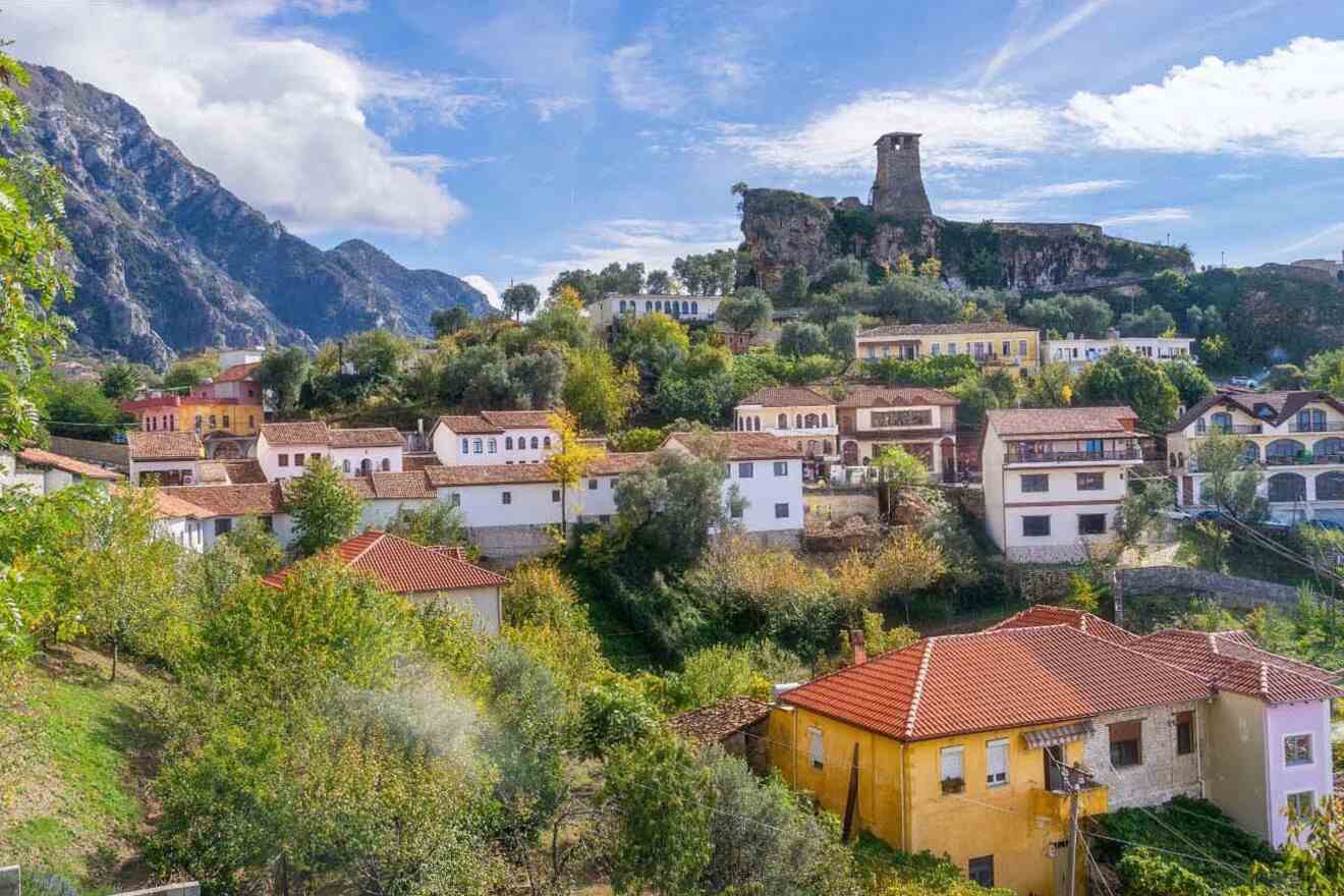 A scenic view of a hillside village with colorful houses, lush greenery, and a historic stone tower atop a hill, set against a mountainous background under a clear sky.