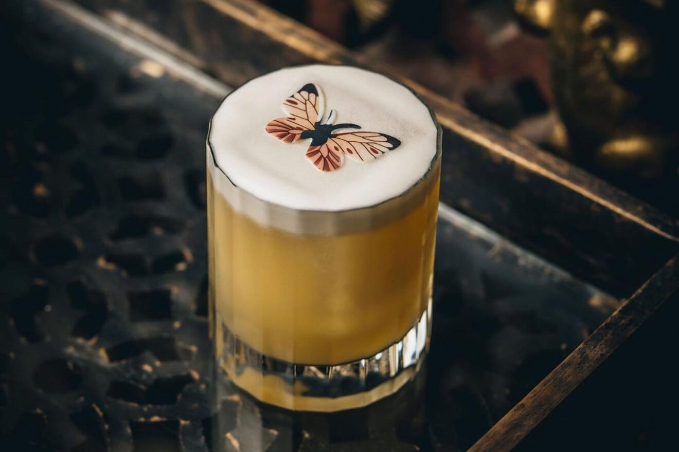 A beautifully crafted cocktail with foam art featuring a butterfly sits on a reflective table, showcasing intricate mixology.