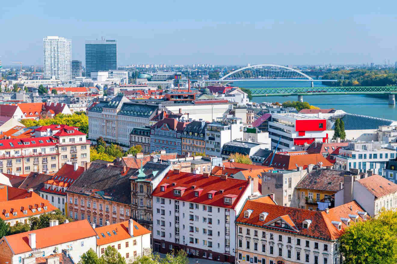 A cityscape shows colorful buildings with red roofs, modern skyscrapers in the background, and a large bridge spanning a river under a clear blue sky.