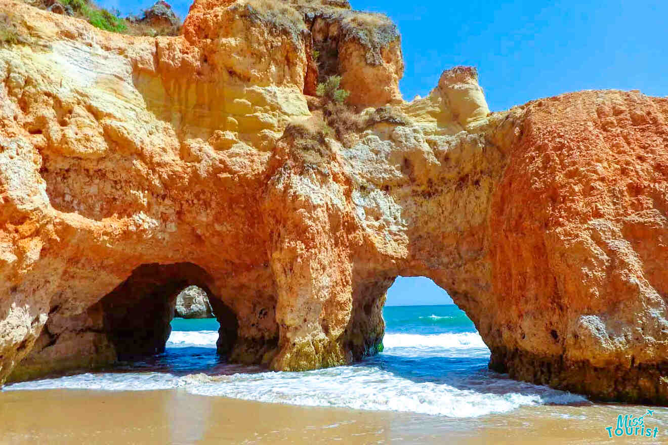 Rocky cliffs with natural arches tower over a sandy beach in Portugal, where waves gently lap the shore. The blue sky and sea are beautifully framed through the arches, creating a stunning coastal scene.