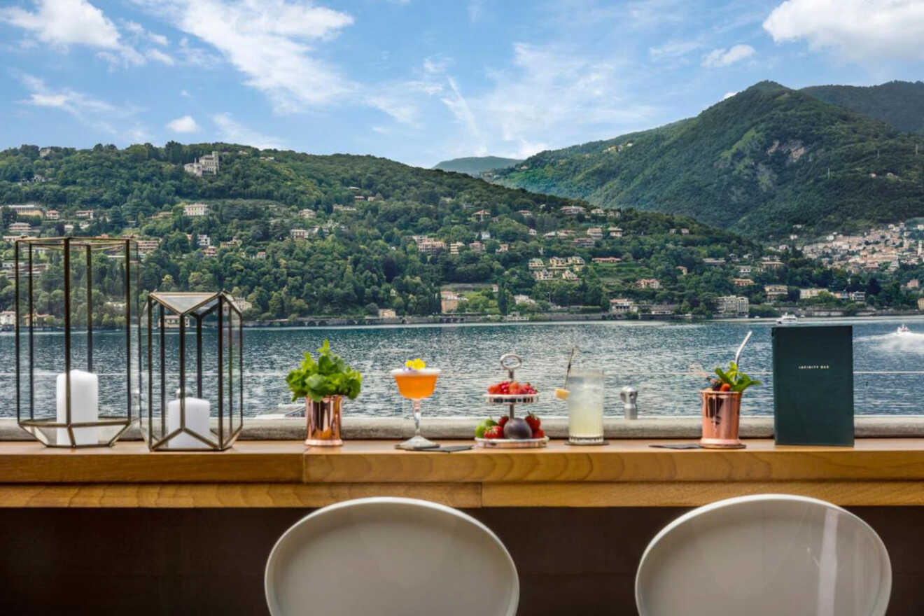 Two white chairs at a wooden table with cocktails, candles, and snacks overlook a serene lake with green hills and houses in the background under a blue, partly cloudy sky.