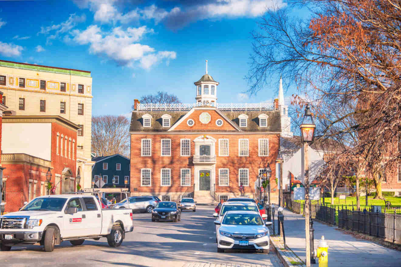 Historic brick building with a cupola, flanked by modern and older structures, with vehicles parked along the street on a sunny day.