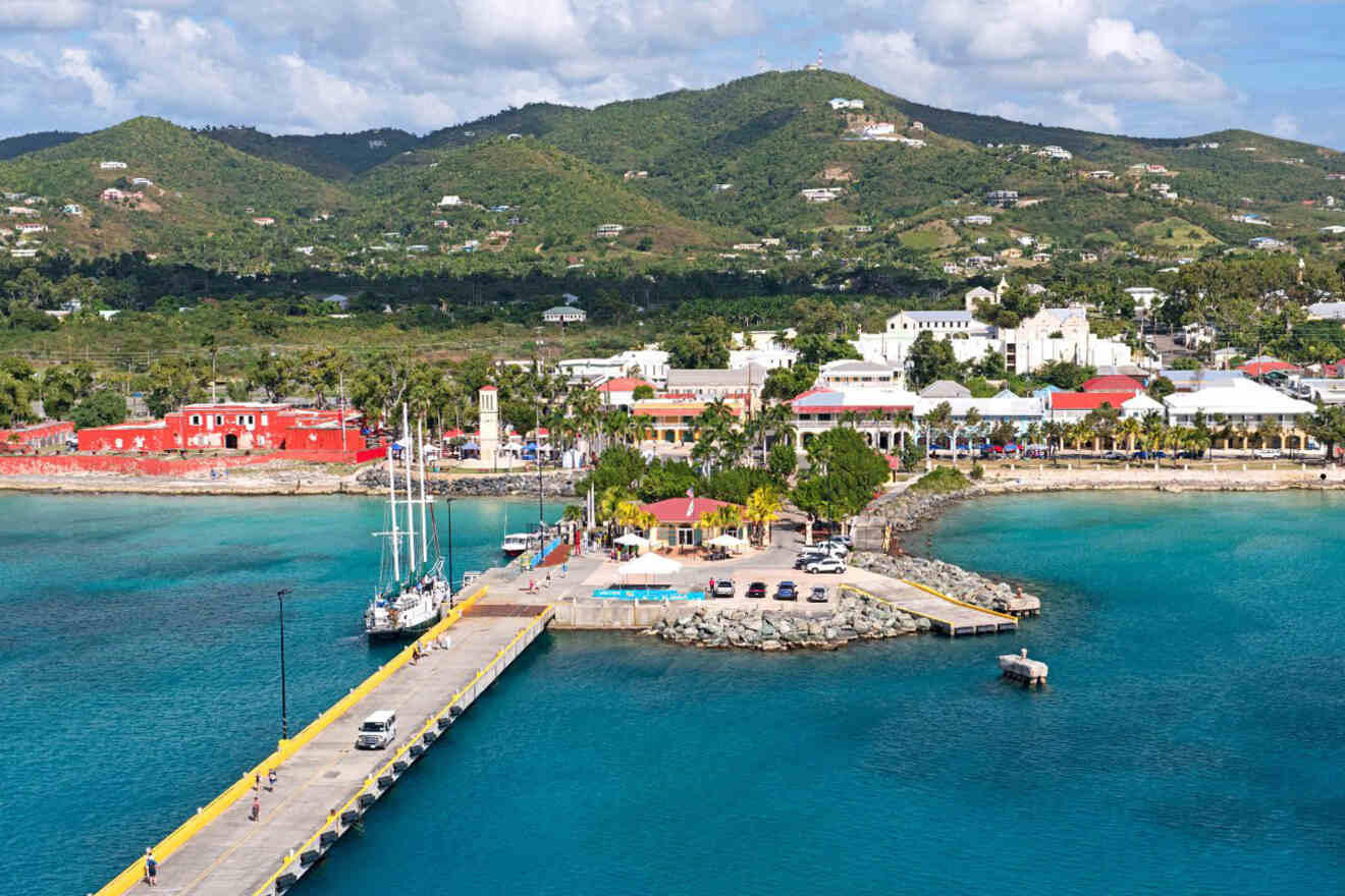 A panoramic view of a coastal town with a dock extending into clear blue water, surrounded by lush green hills and various buildings, including a large red structure and white-roofed buildings.