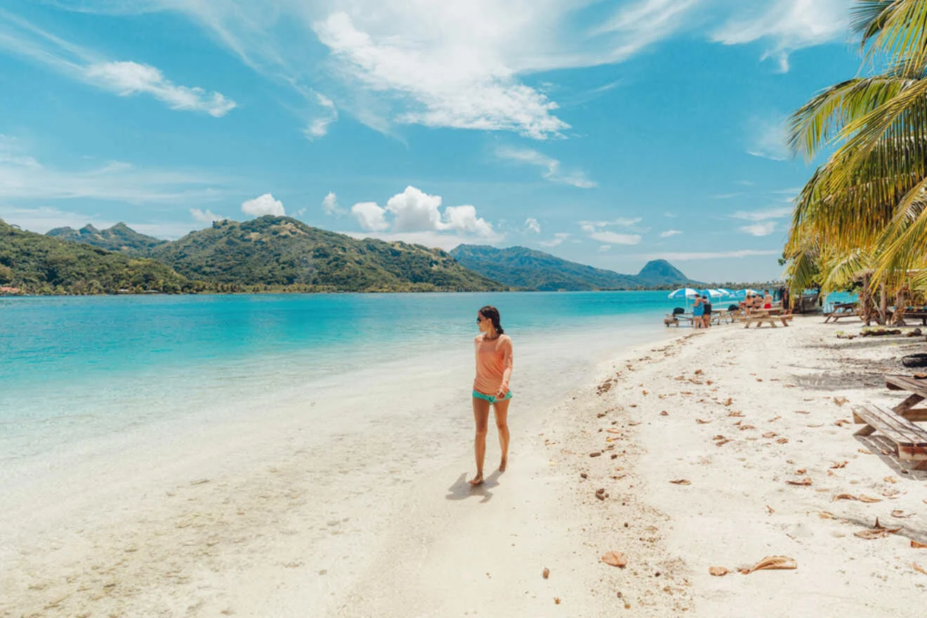 A person walks along a sandy beach with turquoise waters and mountains in the background. There are palm trees and beach chairs to the right.