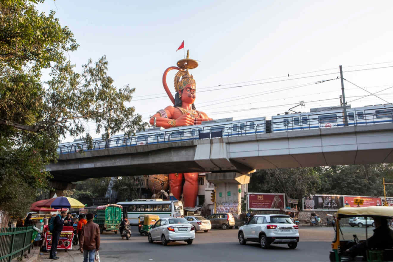 A giant statue of Lord Hanuman near a metro bridge, with vehicles and pedestrians passing underneath in Karol Bagh.