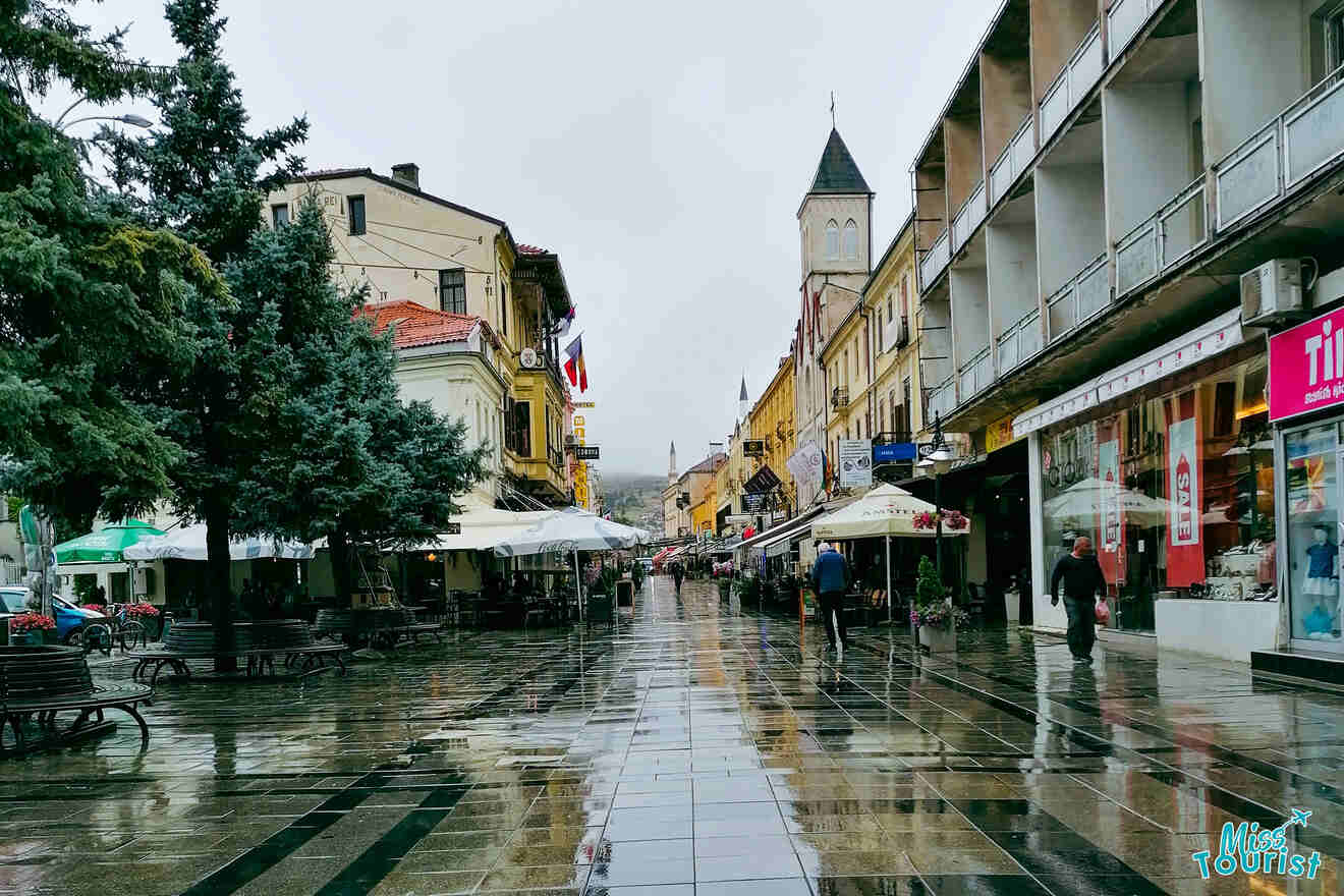 A wet cobblestone street in an urban area lined with shops and outdoor cafe seating. A church tower is visible in the distance under a cloudy sky.