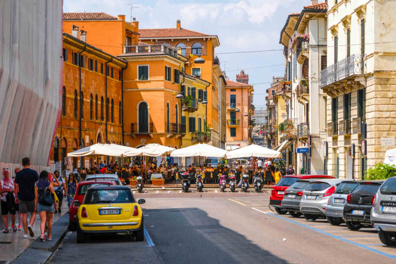 A vibrant street scene shows parked cars, motorcycles, pedestrians, and outdoor dining setups with umbrellas. Buildings in warm hues line both sides of the road under a partly cloudy sky.