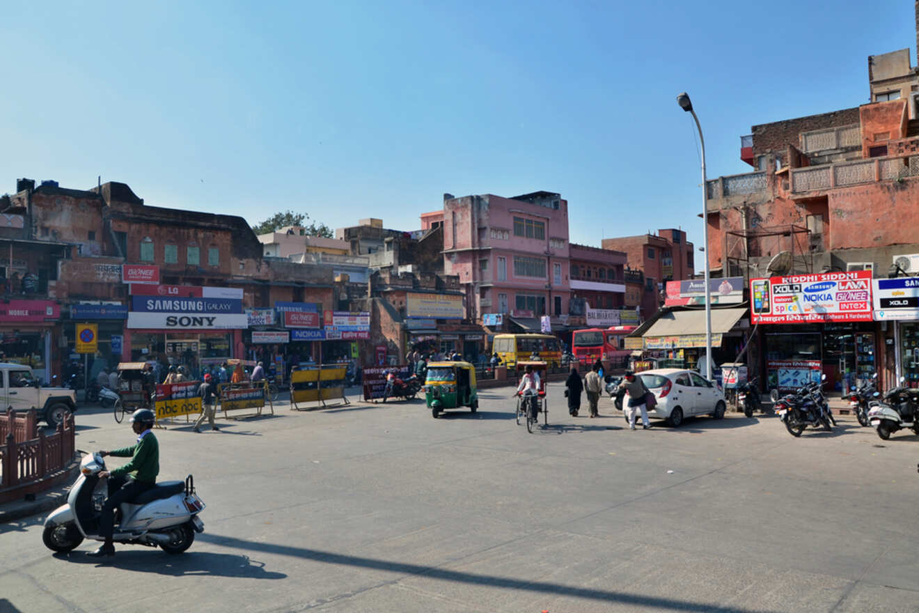 A busy street scene in an urban area with people walking, vehicles, and storefronts. There are various shops and buildings, some in disrepair. Motorbikes and auto-rickshaws are present on the street.