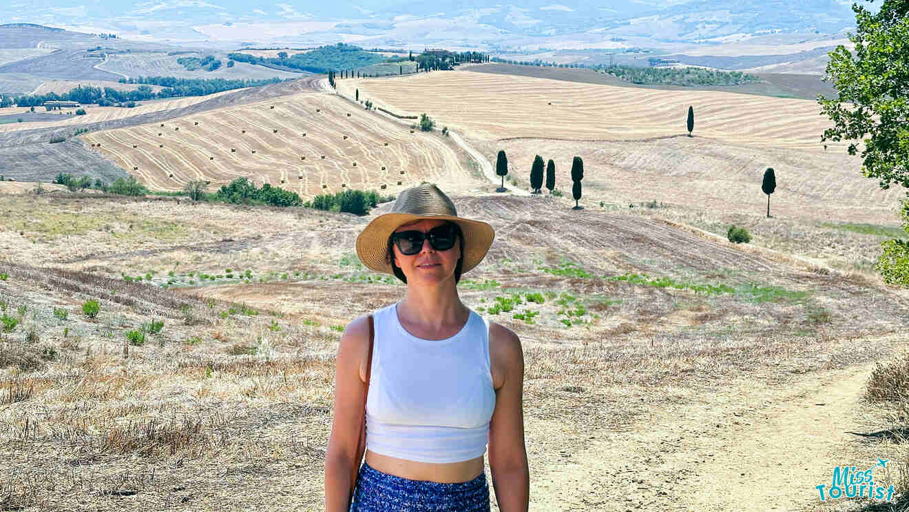 The writer of the post stands in a Tuscan field under a bright sky, wearing a wide-brimmed hat and casual summer outfit, with rolling hills and cypress trees in the background.