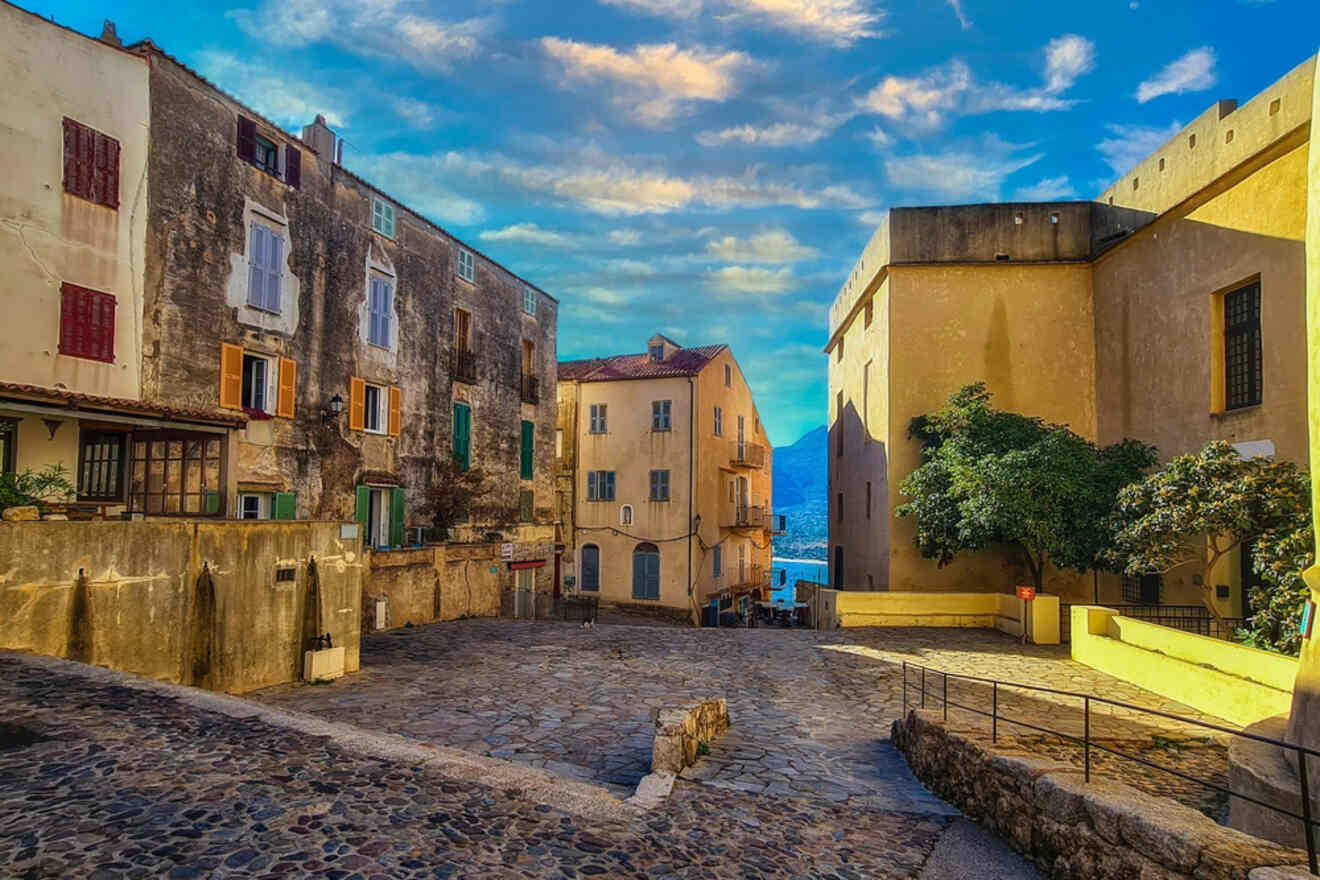 A quiet cobblestone street lined with old buildings featuring colorful shutters and a backdrop of mountains and sea under a dramatic sky.