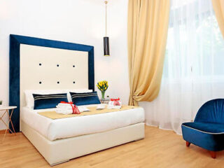 A modern bedroom with a white bed, blue headboard, and matching chair. The bed is decorated with folded towels and flowers. There is a window with beige curtains, and a small side table.