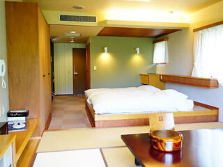 A minimalist hotel room with a wooden bed platform, a white duvet, wooden furniture, and a low table on a tatami mat floor. A doorway and phone are visible in the background.