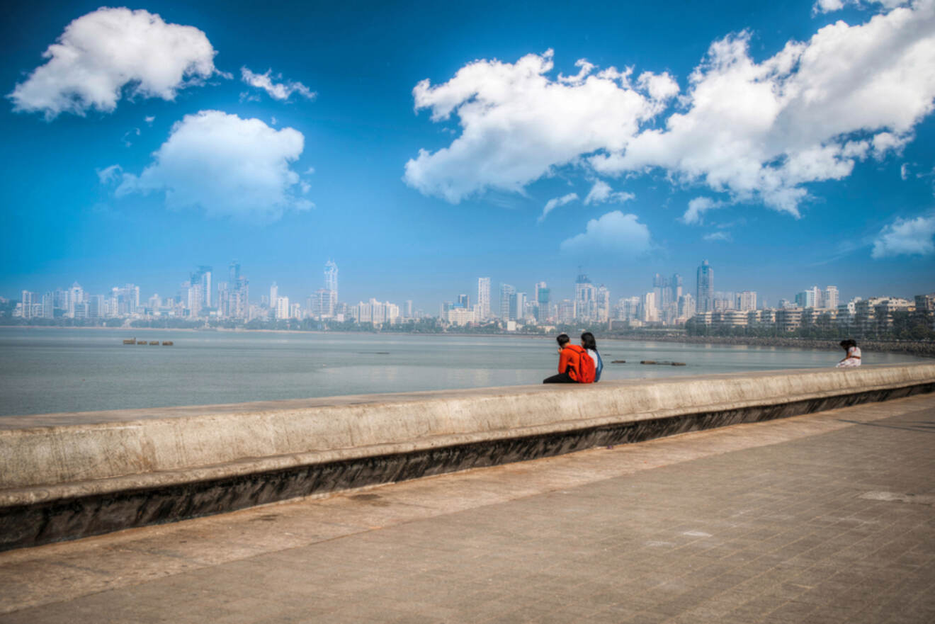 People sit on a seawall overlooking a body of water with a city skyline in the distance under a partly cloudy blue sky.