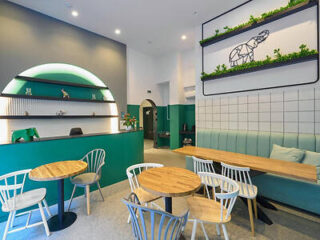 Modern café interior with green and white walls, wooden tables, and chairs. A wall features a plant display and geometric animal art.