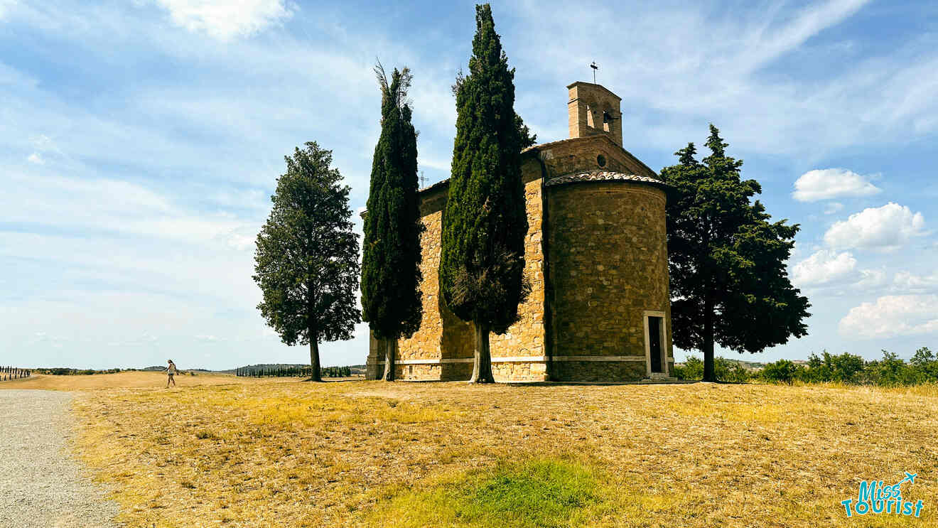 Small, old stone chapel surrounded by tall cypress trees under a clear blue sky in the Tuscan countryside.