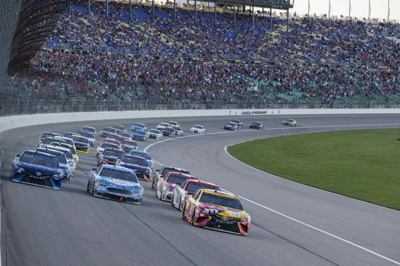 A group of NASCAR cars racing around the track at Kansas Speedway, with the grandstands filled with spectators in the background.