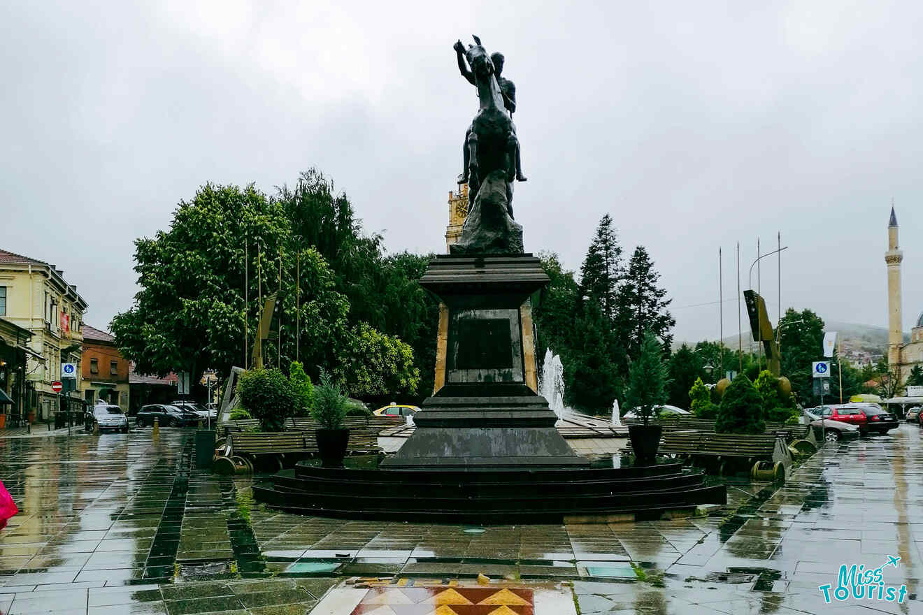 A statue of a man on horseback stands in a city square with wet pavement, surrounded by trees, buildings, benches, and a mosque with a minaret in the background.